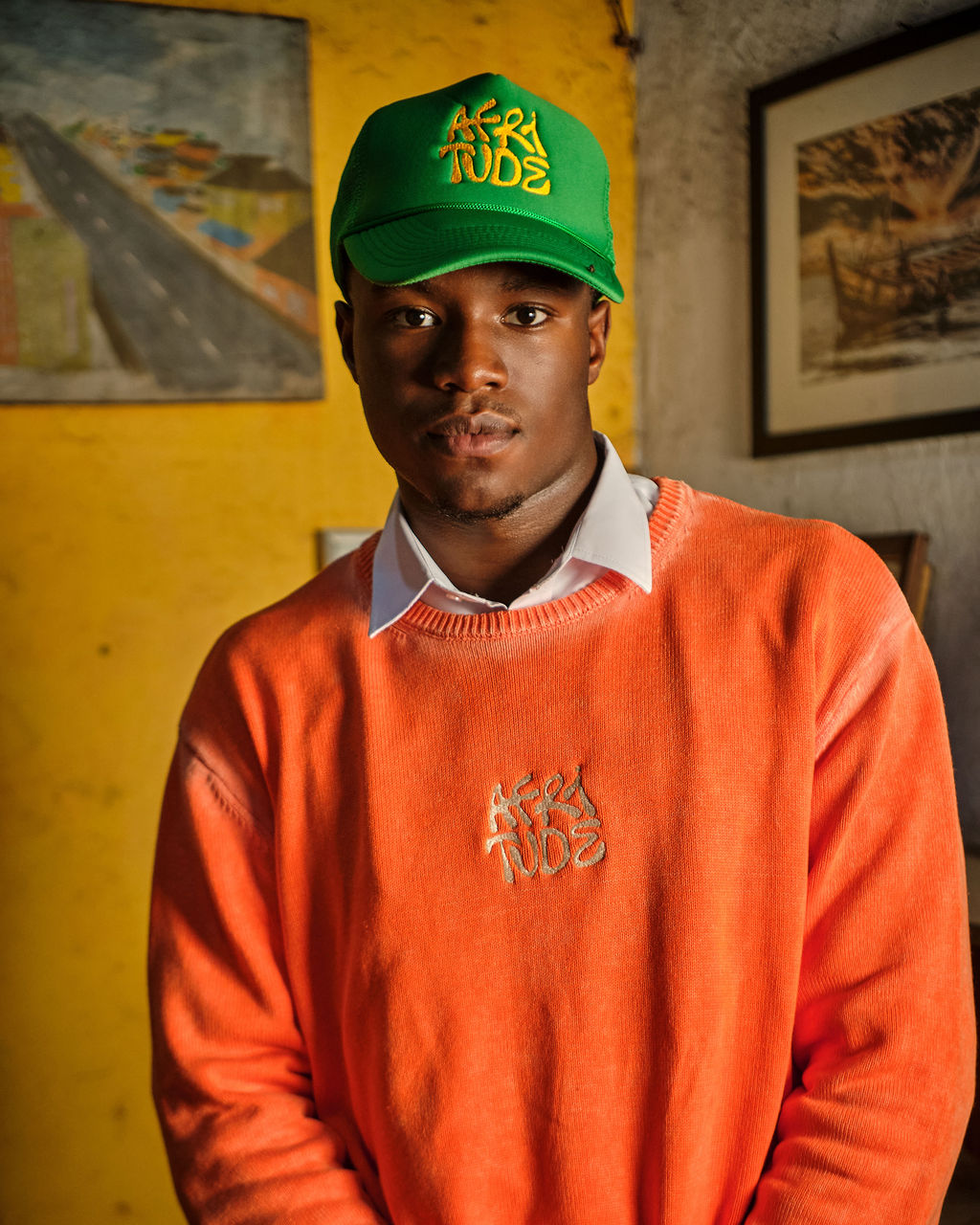 Person wearing a green cap and orange sweater with branding, standing against a yellow wall with framed pictures.